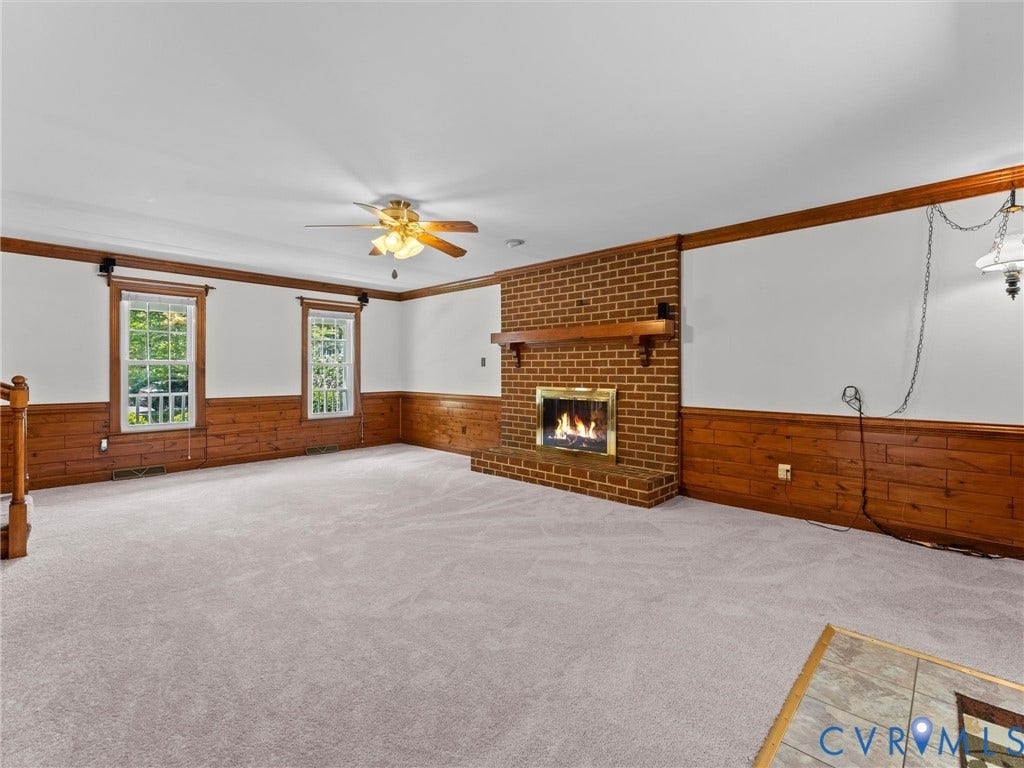 Spacious room featuring a brick fireplace with a wood mantel, light-toned carpeting, wood paneling, and two large windows