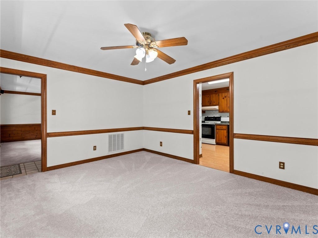 Carpeted interior space featuring white walls with wood trim and chair rail molding, a ceiling fan with light fixture, and doorways to additional rooms