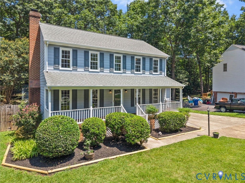 Two-story residence featuring light blue siding with dark shutters, a covered front porch with white railings, a prominent brick chimney, and a concrete driveway