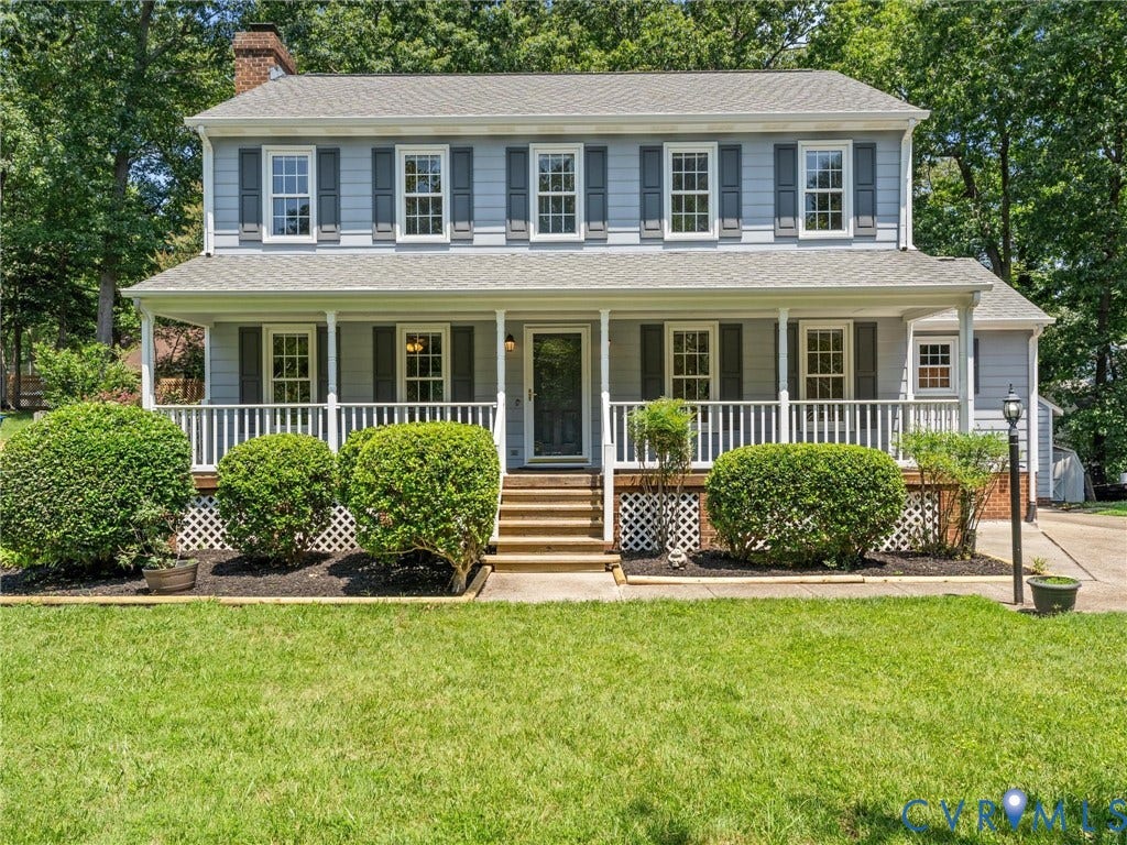 Traditional two-story residence featuring light blue siding with dark shutters, a covered front porch with white railings, a brick chimney, mature front landscaping, and a green lawn