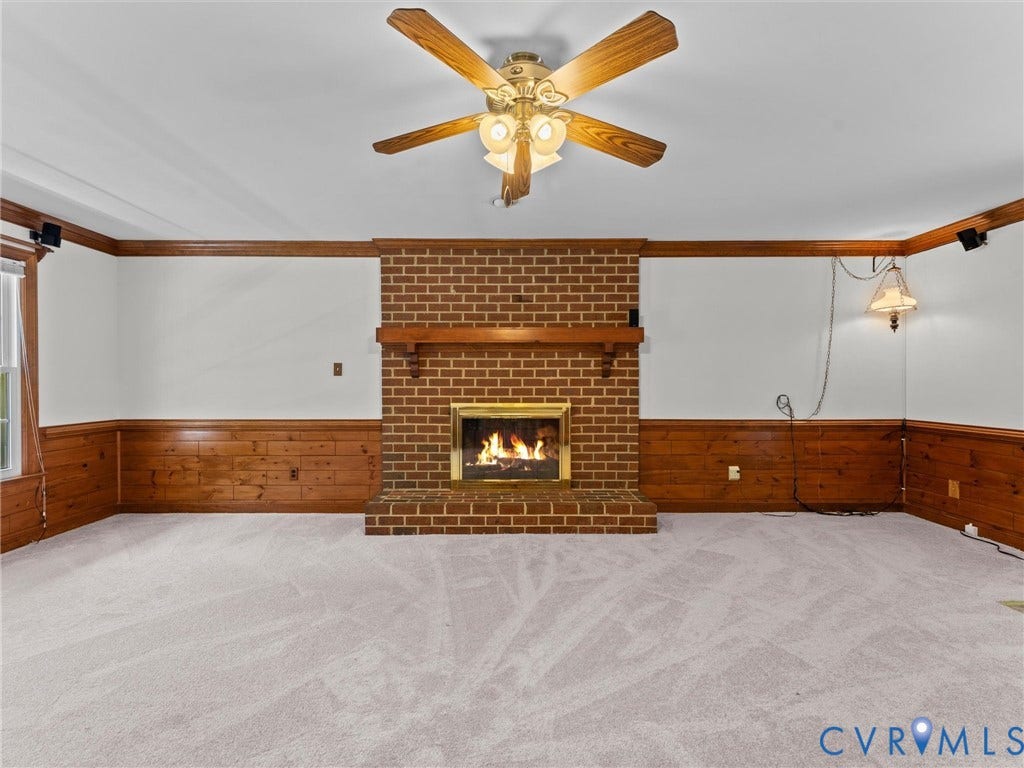 Brick fireplace with a wood mantel, light-toned carpeting, wood wainscoting, white walls, and a ceiling fan with light fixture
