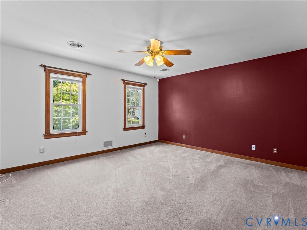Spacious room featuring light gray carpeting, two windows with wood trim, a rich red accent wall, and a ceiling fan with light fixture
