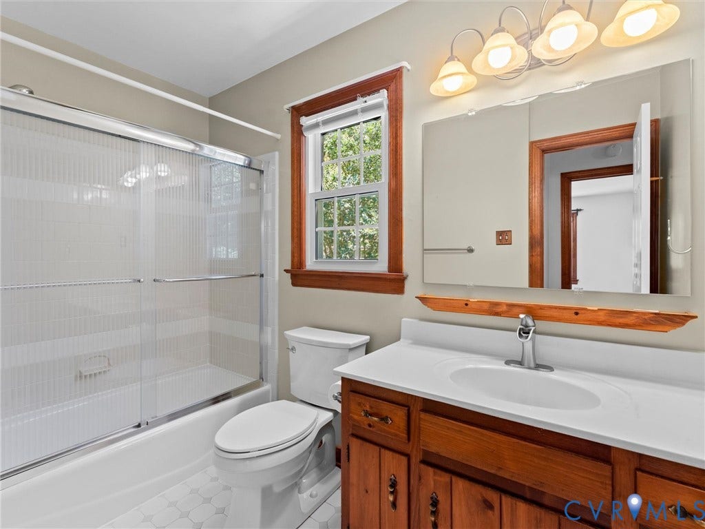 Bathroom featuring a tub/shower combination with sliding glass doors, a vanity with a white countertop and wood-finish cabinetry, a single window with wood-finish trim, and hexagonal floor tiling