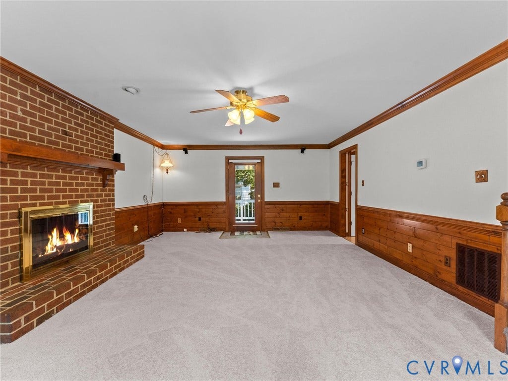 Spacious room featuring a brick fireplace with a brass firebox, light-colored carpeting, wood paneling, and a ceiling fan
