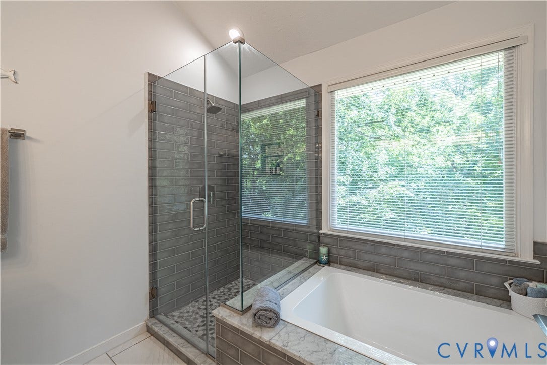 Spacious bathroom featuring a glass-enclosed shower with subway tile surround and pebble flooring, a built-in soaking tub with a tiled base, and a large window with blinds