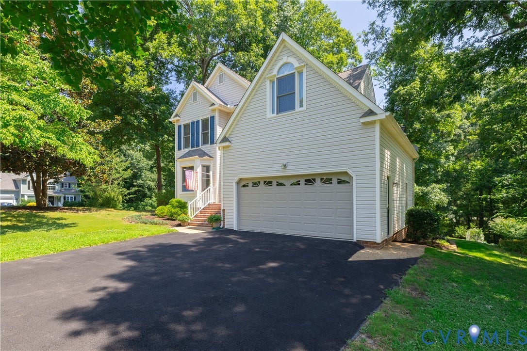 Traditional-style residence featuring light-colored siding, a two-car garage with paneling, a paved driveway, and mature landscaping