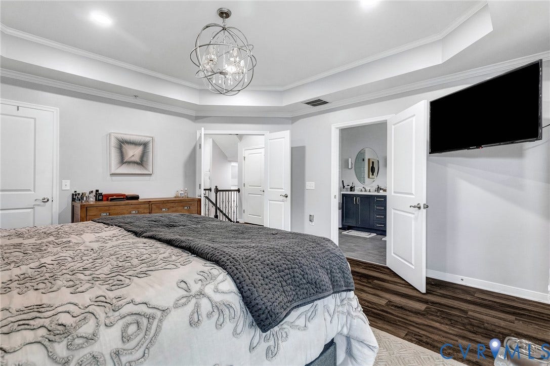 Bedroom featuring a tray ceiling with a contemporary chandelier, wood-finish flooring, and a doorway opening to a bathroom with a modern vanity