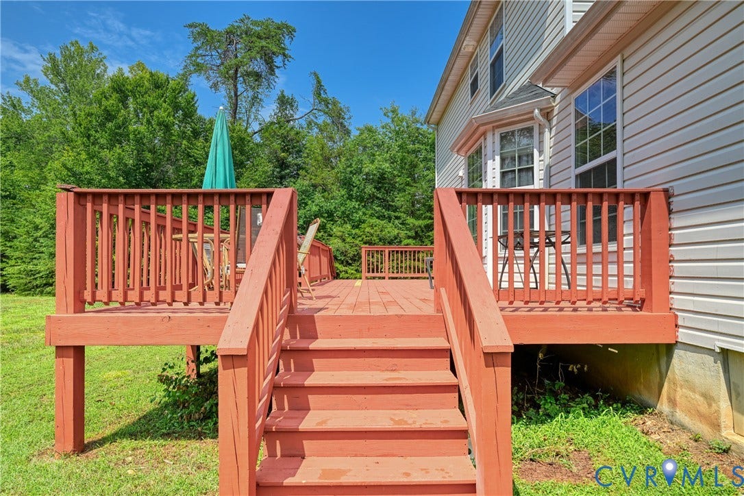 519 N Timber Tribe, Lake Anna, Mineral, VA 23117 Expansive wooden deck with railing and stairs, featuring a light-colored vinyl siding exterior, multiple windows, and a surrounding natural treeline