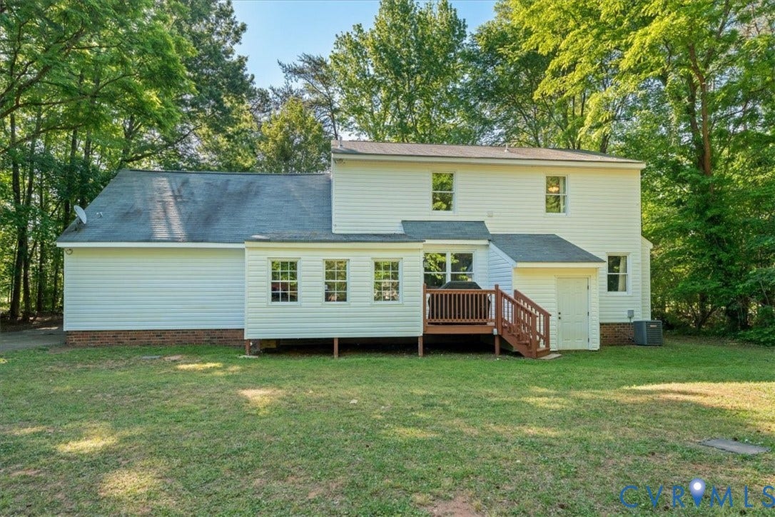 Rear exterior featuring white vinyl siding, a multi-level roofline, a wooden deck with stairs, multiple windows, and a mature treed backdrop