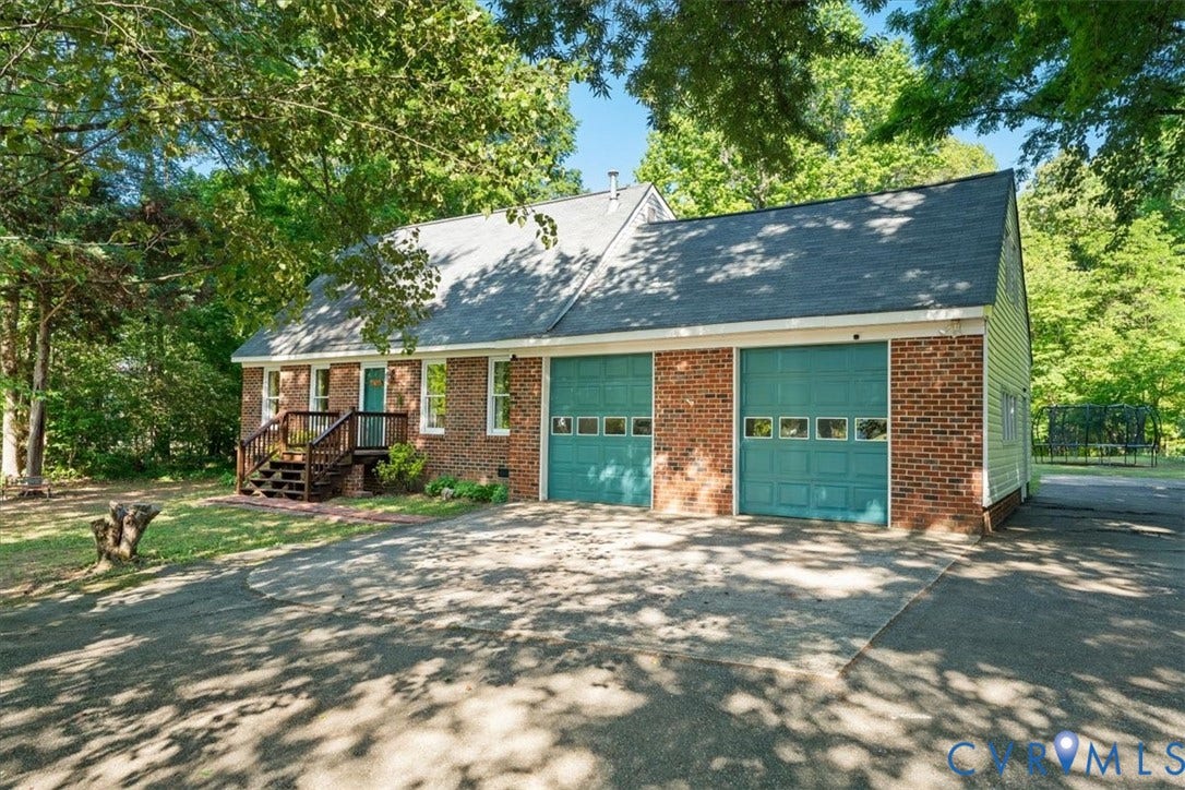Brick exterior featuring a two-car garage with teal doors, a shingled roof, a front porch with wooden steps and railing, and surrounding mature trees