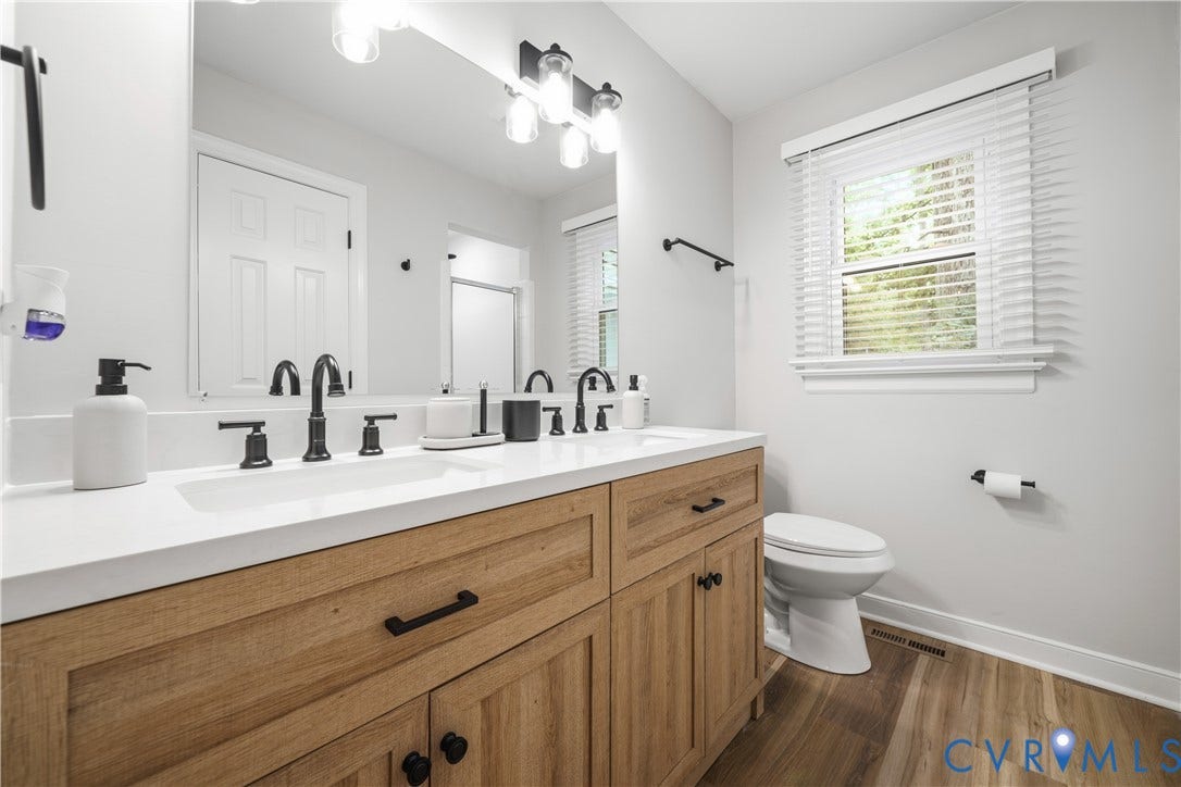 Double vanity with a white countertop and integrated sinks, wood-finish cabinetry with black hardware, matte black faucets, and overhead vanity lighting