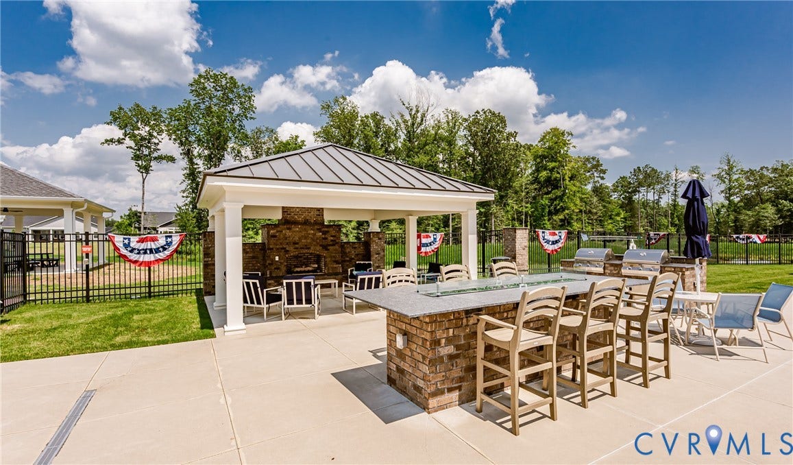 7627 E Danube Loop, New Kent, VA 23124 Outdoor living area featuring a covered gazebo with a stone-clad fireplace, a large brick-base bar with a light-toned countertop, multiple built-in grills, and extensive concrete patio space surrounded by a black metal fence