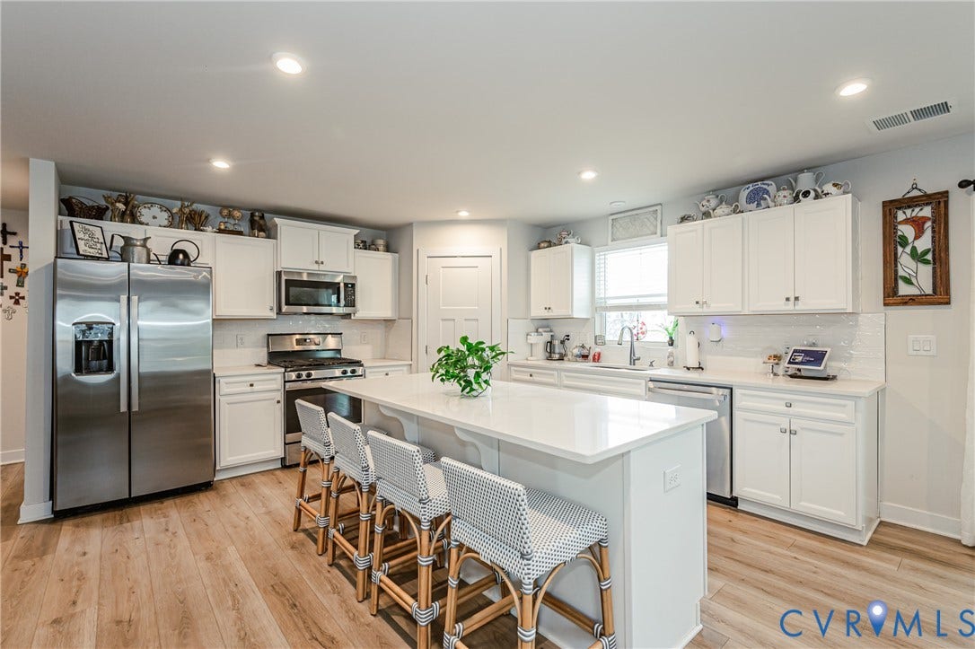 7627 E Danube Loop, New Kent, VA 23124 Modern kitchen featuring a large central island with seating, white shaker-style cabinetry, stainless steel appliances, wood-finish flooring, and recessed lighting