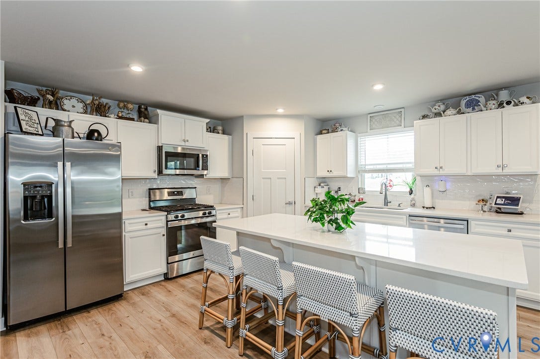 7627 E Danube Loop, New Kent, VA 23124 Bright kitchen featuring a central island with a solid surface countertop, white cabinetry, light wood-finish flooring, stainless steel appliances, and a tiled backsplash