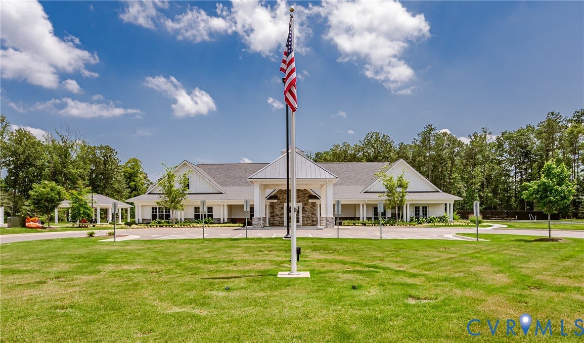 7627 E Danube Loop, New Kent, VA 23124 Expansive lawn with a central flagpole, bordered by a long white-sided building featuring stone accents, multiple gables, and a columned front porch