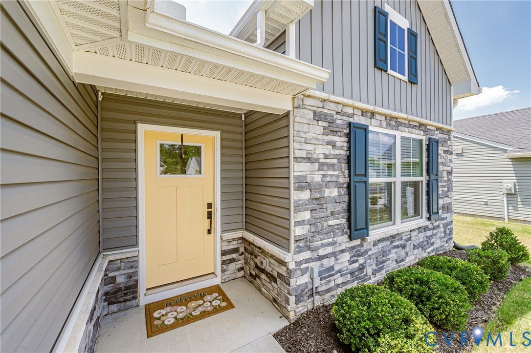 7627 E Danube Loop, New Kent, VA 23124 Exterior featuring a light yellow entry door with a window pane, a covered concrete porch, grey horizontal siding, grey vertical siding, and a stacked stone facade