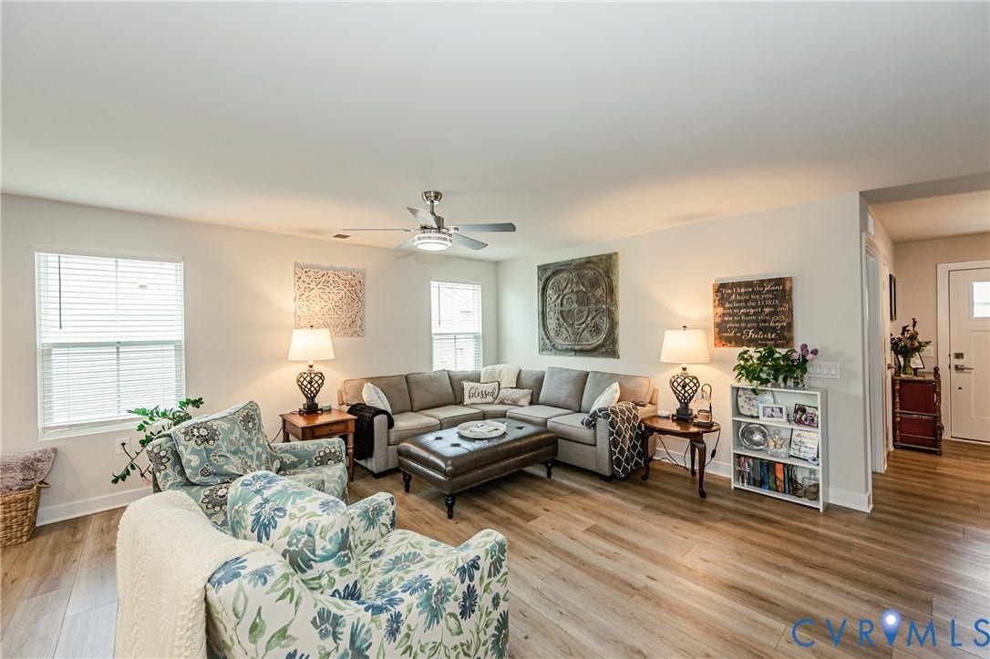 7627 E Danube Loop, New Kent, VA 23124 Spacious living room featuring wood-finish flooring, white walls, multiple windows with blinds, and a ceiling fan with integrated lighting