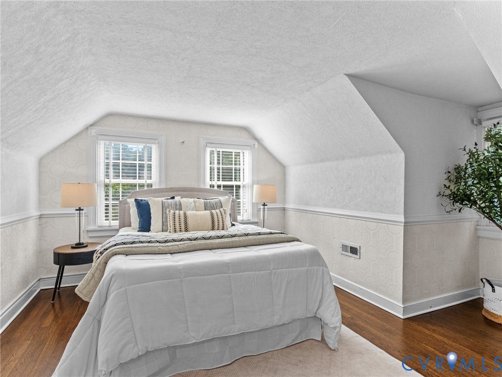 Hardwood flooring with a light area rug, two double-hung windows with blinds, and white wainscoting with a subtle patterned wallpaper