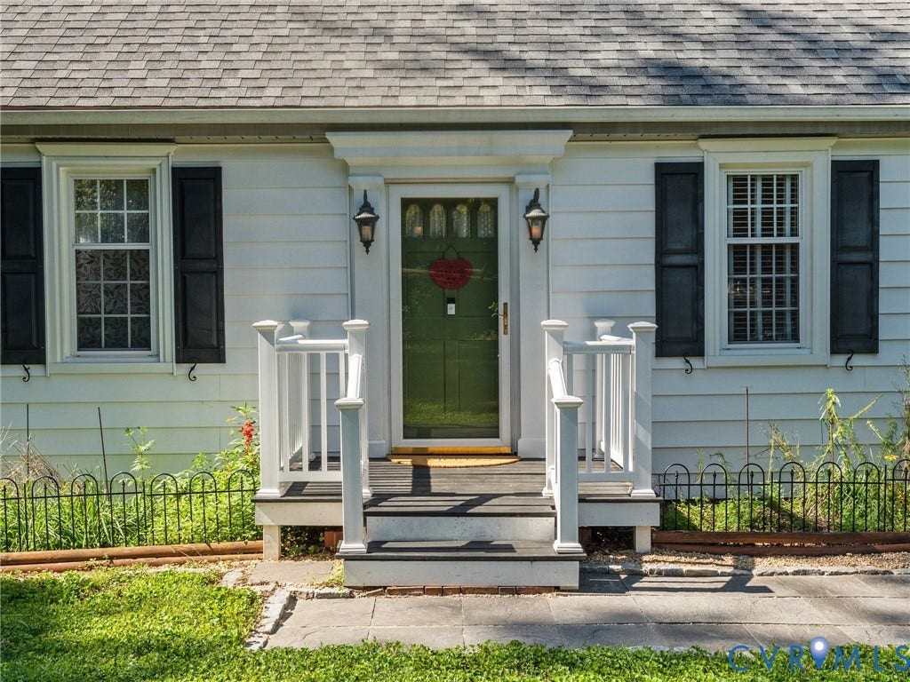 Charming white exterior with a green entry door, featuring decorative black shutters, white window trim, a gray shingle roof, and a welcoming porch with white railings