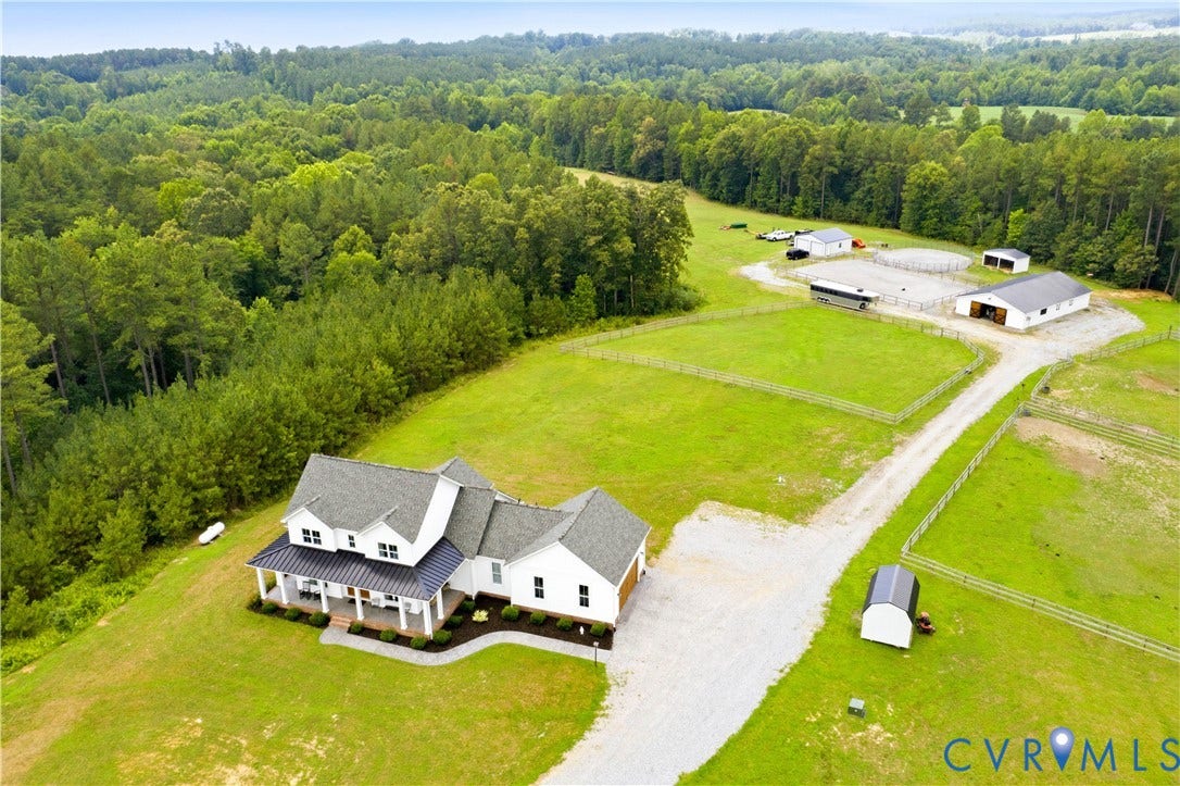25181 W Soap Stone Rd, Amelia, VA 23083 Expansive property featuring a white two-story residence with a covered front porch, gravel driveway, multiple outbuildings, fenced pastures, and a circular riding arena, all surrounded by mature woodlands