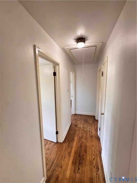 Hallway featuring wood-finish flooring, light-toned walls, multiple doorways, a ceiling-mounted light fixture, and an attic access panel