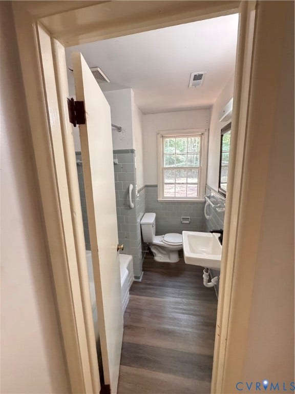 Full bathroom featuring wood-finish flooring, light blue tiled walls, a window with exterior views, a wall-mounted sink, and a commode