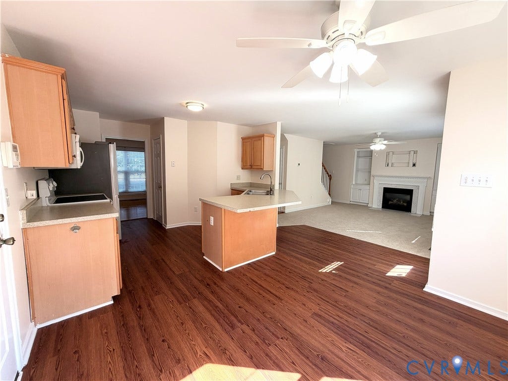 Wood-finish flooring, light-toned cabinetry, a kitchen island with sink, ceiling fans, and a living area with a fireplace