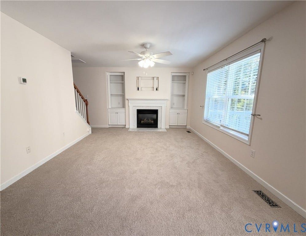 Living space featuring carpeting, a fireplace with a white mantel, built-in shelving, a ceiling fan, and a large window