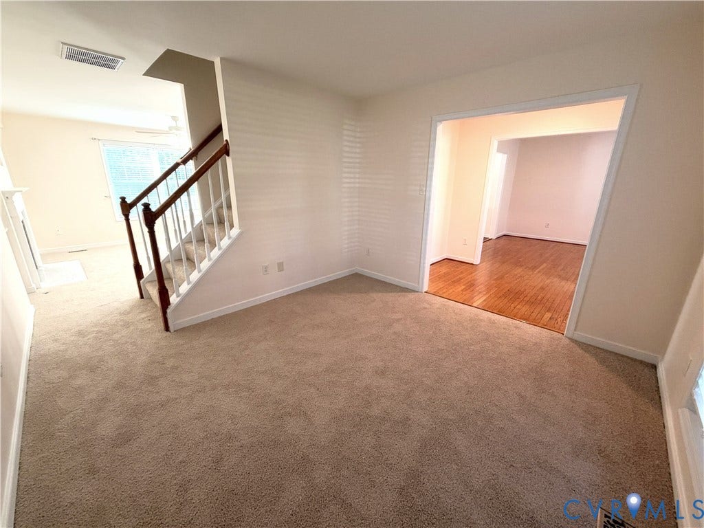 Neutral-toned carpeting, staircase with wood banister and white spindles, hardwood flooring in adjacent room, white trim throughout, and a window with blinds