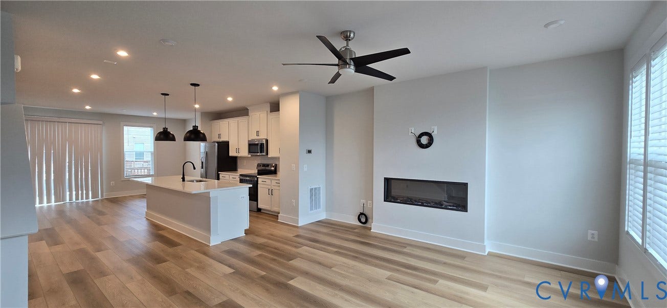 Open-concept living area featuring wood-finish flooring throughout, a modern kitchen with white cabinetry and stainless steel appliances, pendant lighting over the island, a ceiling fan, and a linear fireplace accent wall with integrated shelving