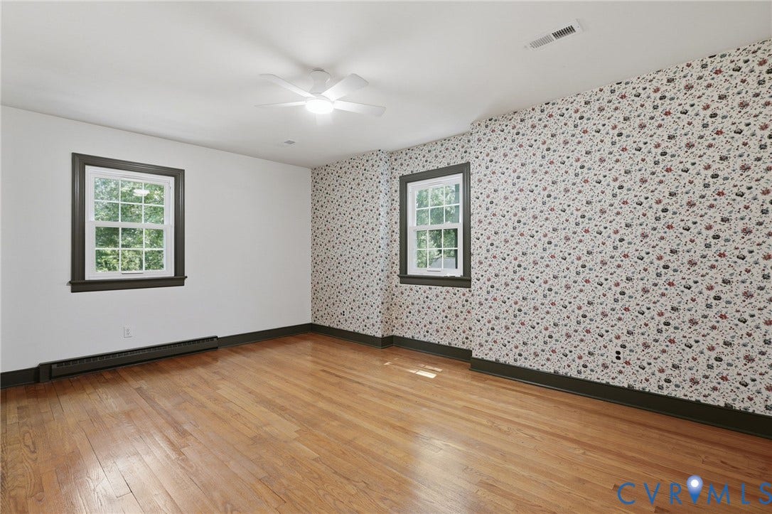 Hardwood flooring throughout featuring two double-hung windows with dark trim, a ceiling fan with integrated lighting, and baseboard heating