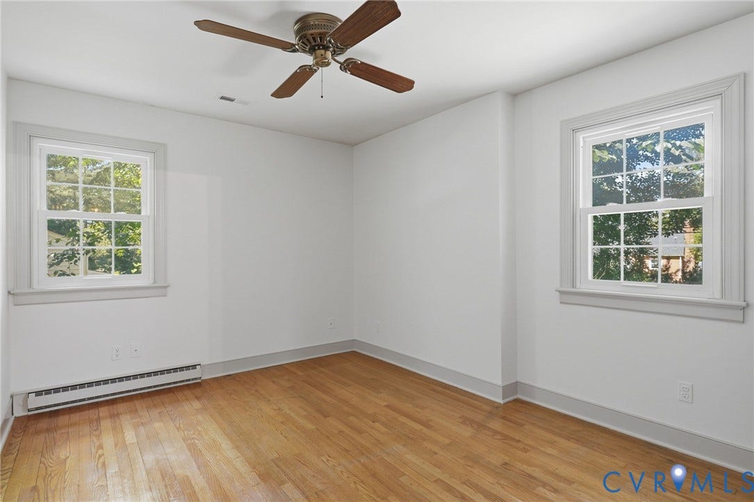 Hardwood flooring throughout this room, featuring two windows with white trim, a ceiling fan with wood-finish blades, and a baseboard heating unit