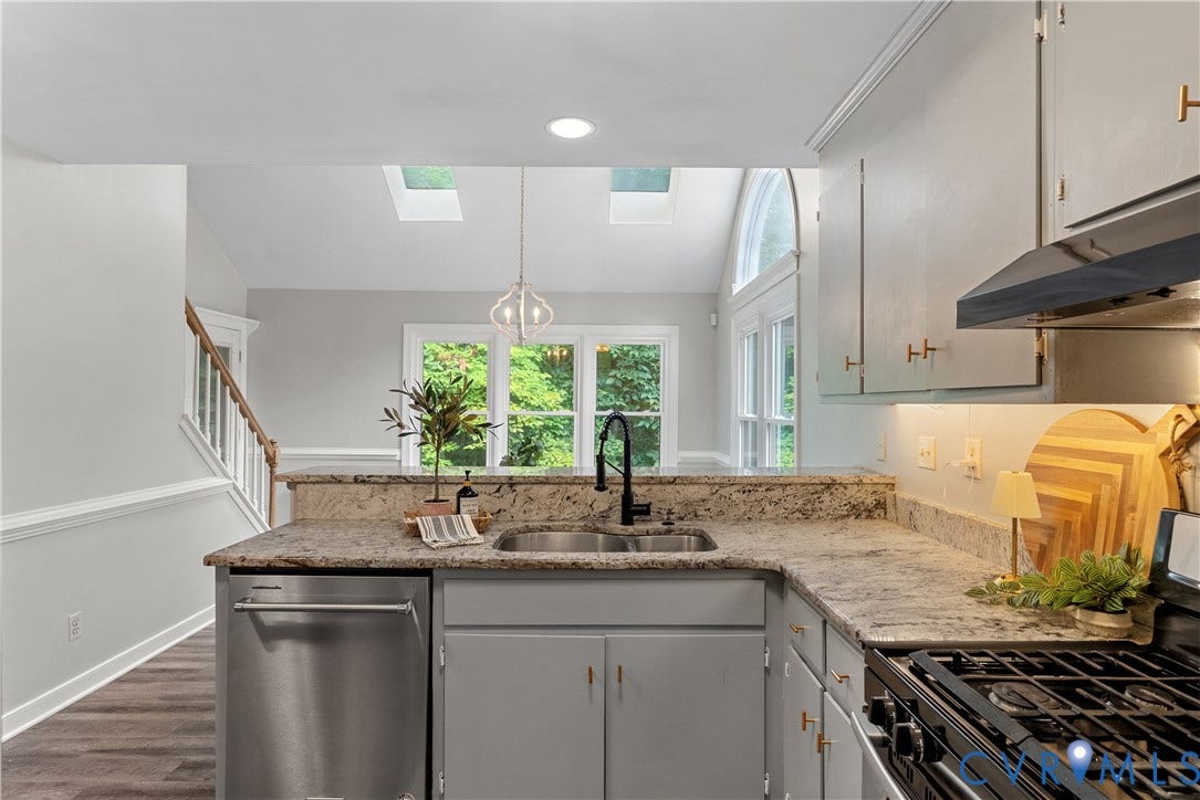 Contemporary kitchen featuring a large granite-topped island with integrated sink and stainless steel dishwasher, professional-grade gas range with stainless steel hood, soft-close cabinetry with brushed brass hardware, recessed and pendant lighting, and skylights flooding the space with natural light