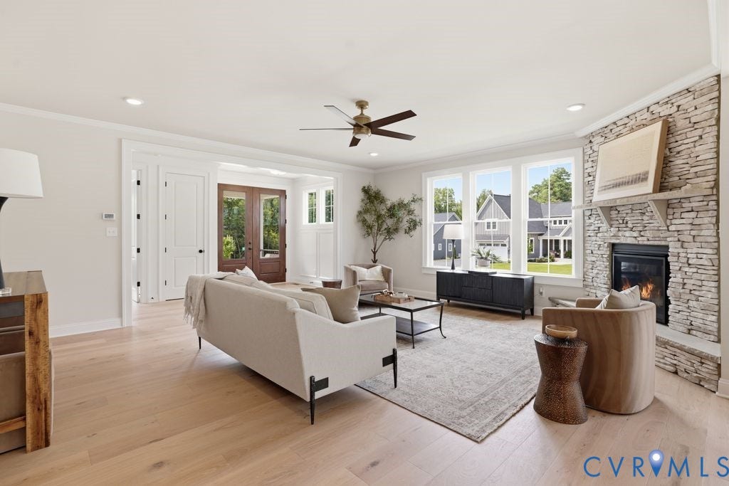Spacious living area featuring light wood-finish flooring, a stone-faced fireplace with hearth and mantle, a ceiling fan, recessed lighting, and multiple windows
