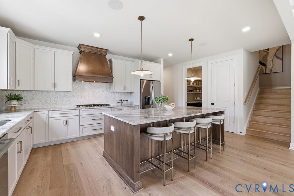 Spacious kitchen featuring an expansive island with seating, light wood-finish flooring, white cabinetry, integrated stainless steel appliances, and a wood-finish range hood