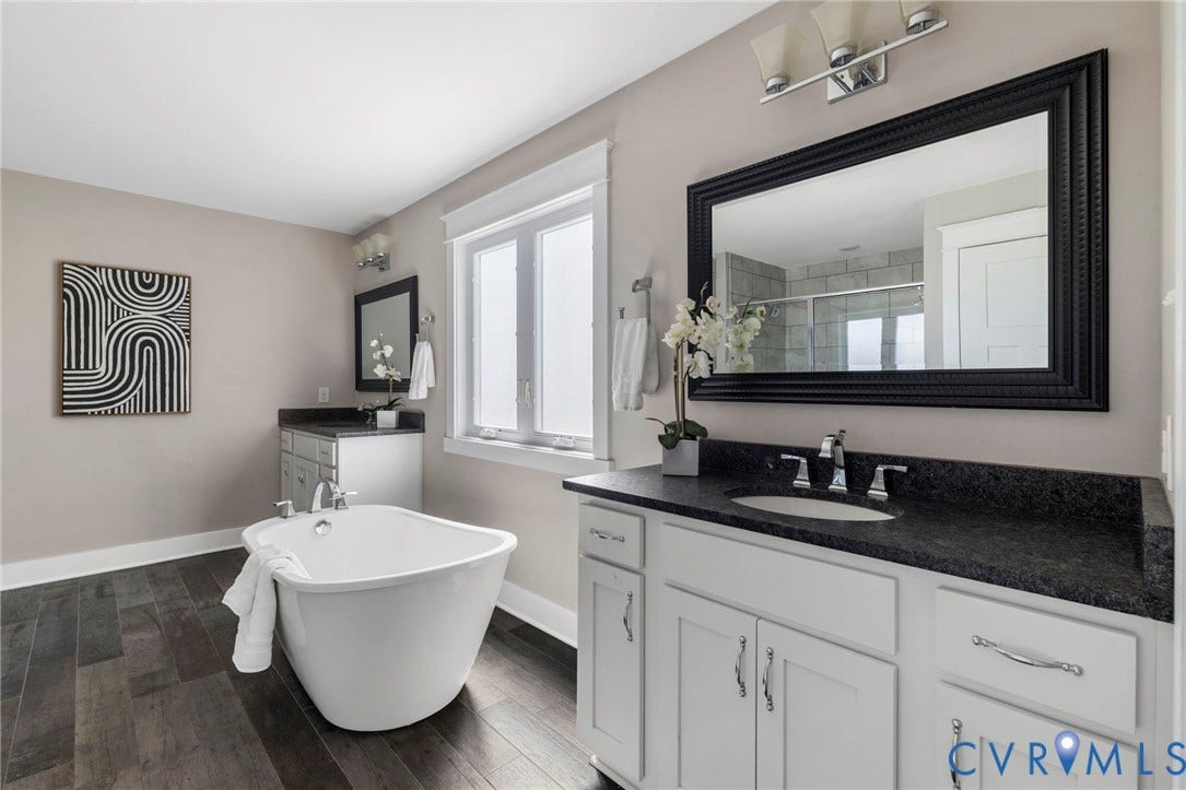 6984 Carden Park Drive, Moseley, VA 23120 Bathroom featuring a freestanding soaking tub, wood-finish flooring, dual white vanities with dark countertops, and a window with privacy glass