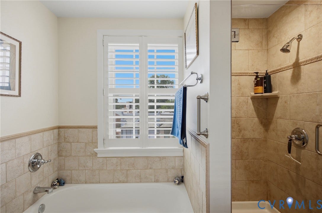 Bathroom featuring a built-in bathtub, a walk-in shower with glass enclosure, light-toned wall tiling, white plantation shutters, and chrome fixtures