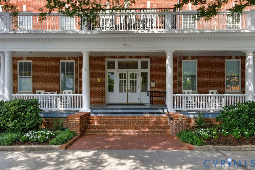 Brick facade building featuring a white columned front porch, white upper-level balcony with railings, white French-style entry doors, and mature landscaping