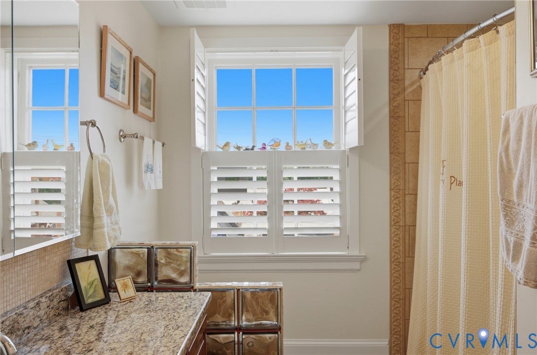 Bathroom featuring a window with white louvered shutters, a granite-finish countertop, and a tiled shower surround