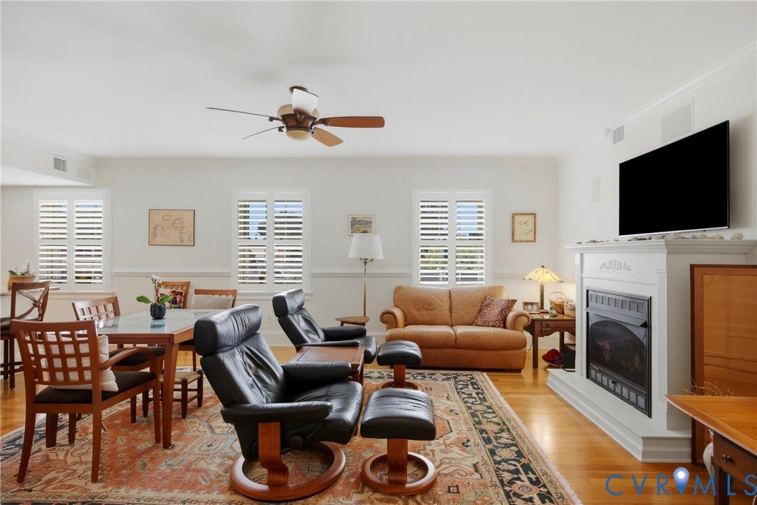 Open-concept living area featuring light wood-finish flooring, white plantation shutters, a ceiling fan, and a fireplace with a white mantel