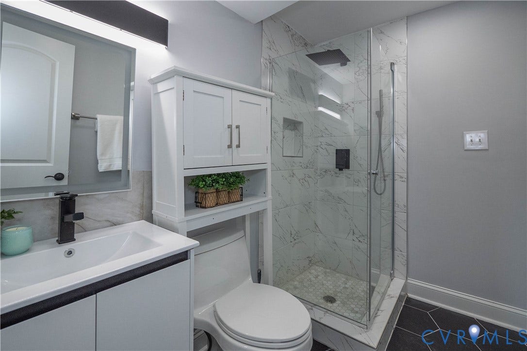 1209 N 22nd Street, Church Hill, Richmond, VA 23223 Contemporary bathroom featuring a glass-enclosed shower with marble-look tile surround and built-in niches, a white vanity with integrated sink and matte black faucet, a mirrored medicine cabinet, an over-the-toilet storage cabinet, and dark hexagonal floor tiles