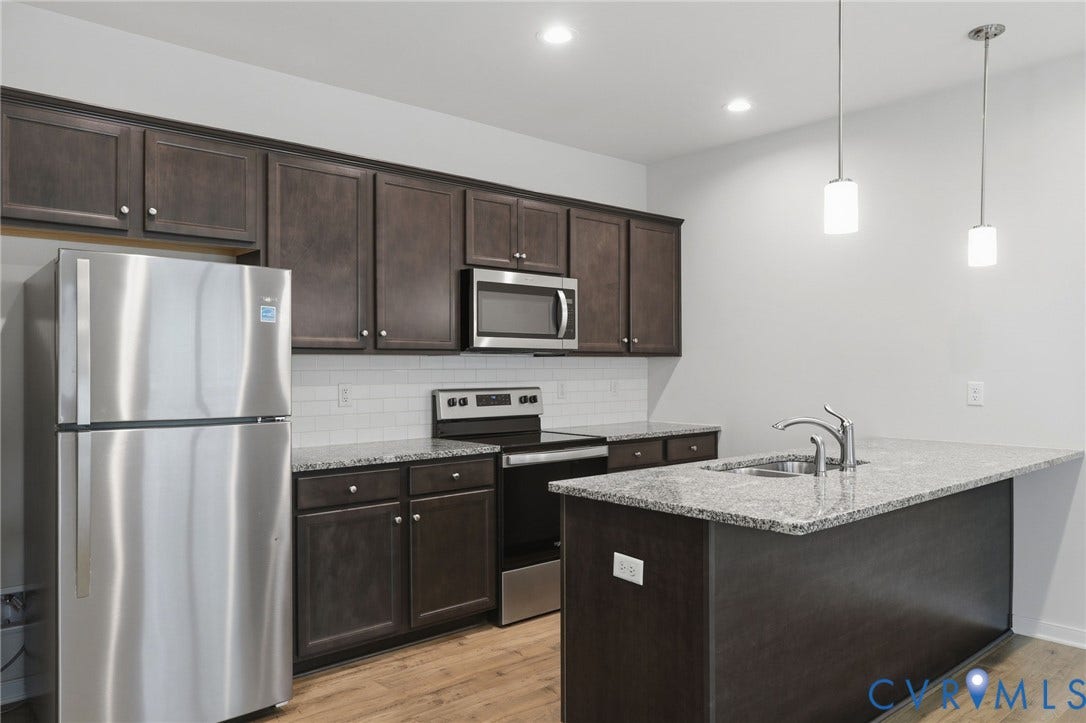 6544 Bolles Landing Court, Chester, VA 23831 Kitchen featuring wood-finish flooring, dark wood cabinetry, stainless steel appliances, a subway tile backsplash, and a kitchen island with a double basin sink and stone countertop