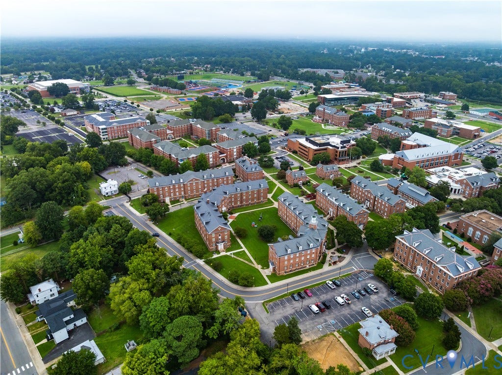 Campus setting featuring multiple brick buildings with dormer windows, extensive green spaces, mature trees, and designated parking areas