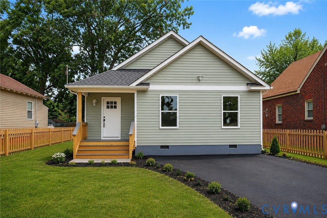 Sage green siding exterior with a dark shingle roof, featuring a covered front porch with natural wood accents, a black asphalt driveway, and a wooden picket fence