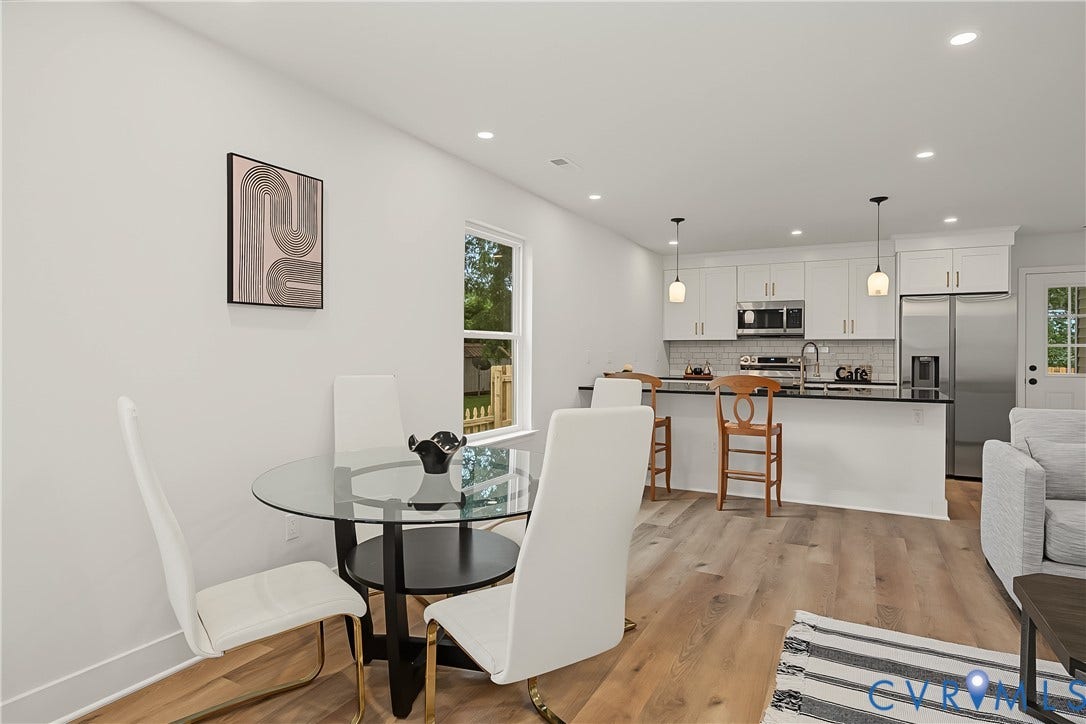 Open-concept dining and kitchen area featuring a round glass-top dining table with white upholstered chairs, wood-finish flooring throughout, and stainless steel appliances