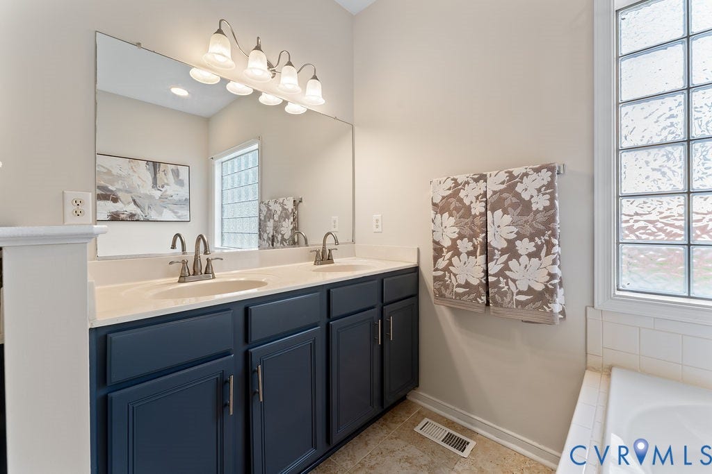 5316 Lemoore Drive, Glen Allen, VA 23059 Bathroom featuring a dual-sink vanity with blue cabinetry and white countertops, a large vanity mirror, a four-light fixture, and a tiled tub surround with a large privacy window