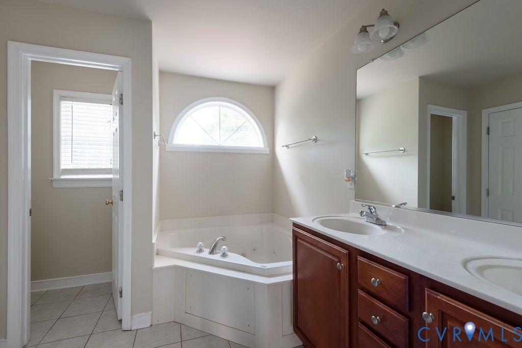 4924 Sadler Glen Ct, Sadler Glen, Glen Allen, VA 23060 Bathroom featuring a corner soaking tub with an arched window, a dual-sink vanity with wood-finish cabinetry and a white countertop, and tiled flooring