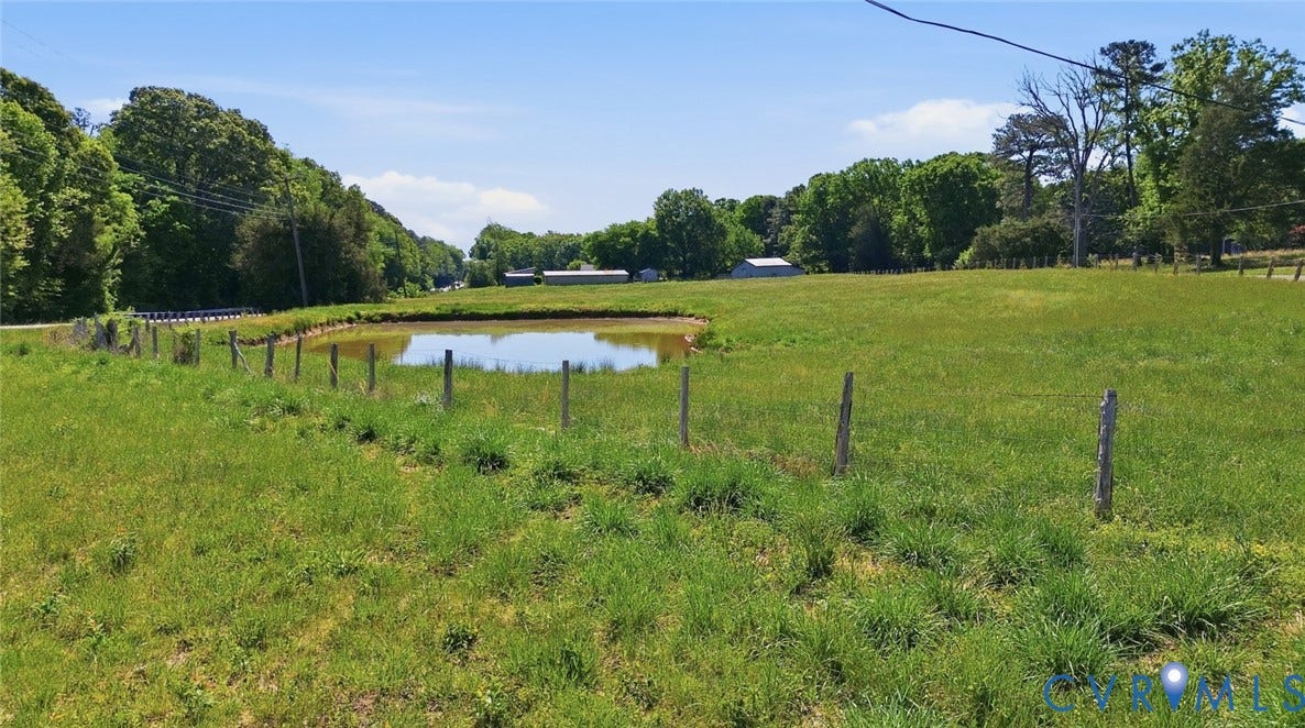 Expansive green field featuring a tranquil pond, mature trees, and a partial perimeter fence