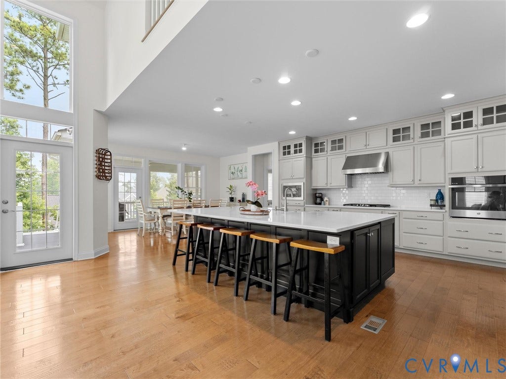 6642 Stonewolf Court, Moseley, VA 23120 Open-concept kitchen featuring a large central island with seating, light wood-finish flooring, white subway tile backsplash, stainless steel appliances, and abundant cabinetry with glass-front accents