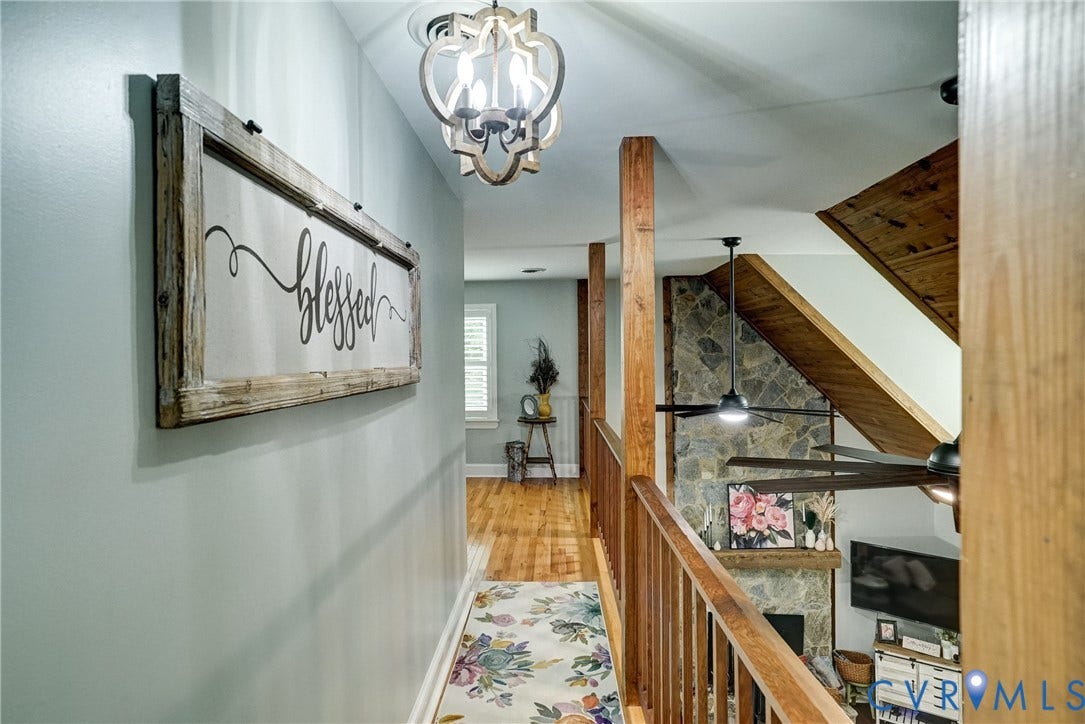 1174 Dorset Road, Powhatan, VA 23139 Hallway featuring wood-finish flooring, a decorative overhead chandelier, and a wood railing overlooking a lower level with a stone-clad fireplace, wood mantel, and wood accent beams